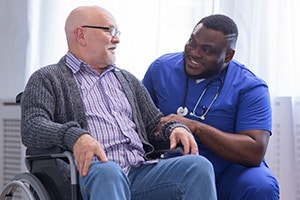 Male nurse kneeling down to speak with a resident seated in a wheelchair.